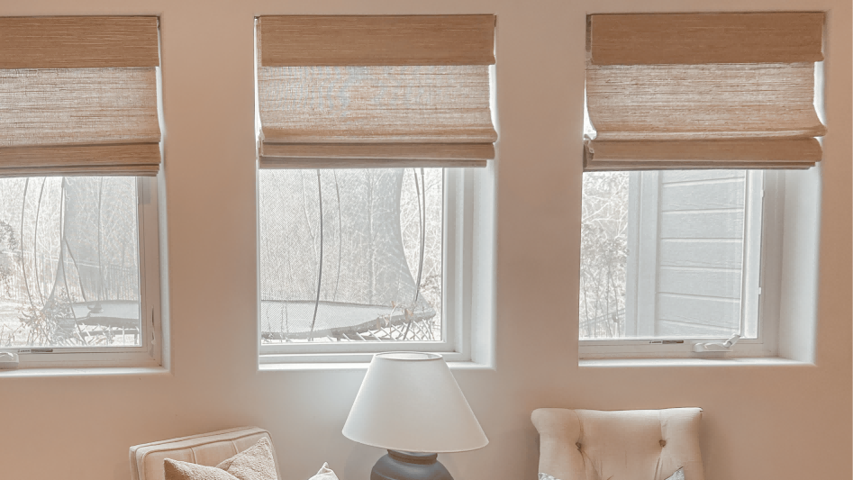 Three light brown bamboo shades partially raised over a series of windows in a bright room, with a lamp and seating area visible below