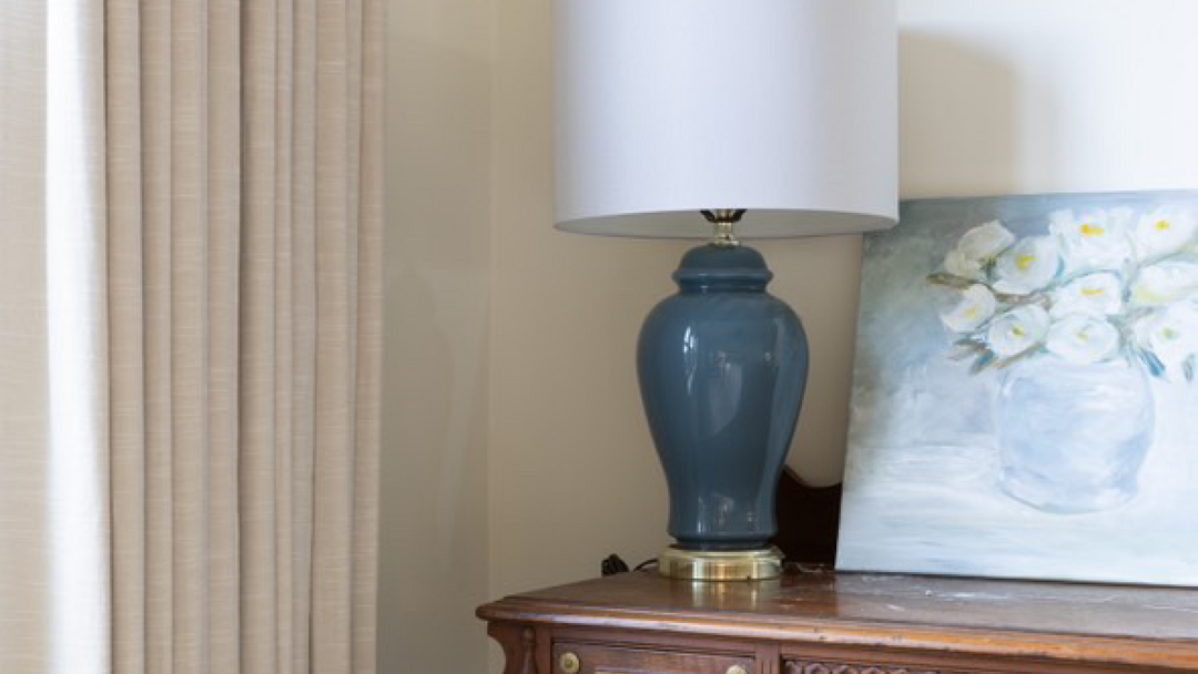 Beige linen curtains frame a window, showcasing a stylishly arranged side table with a blue lamp and artwork